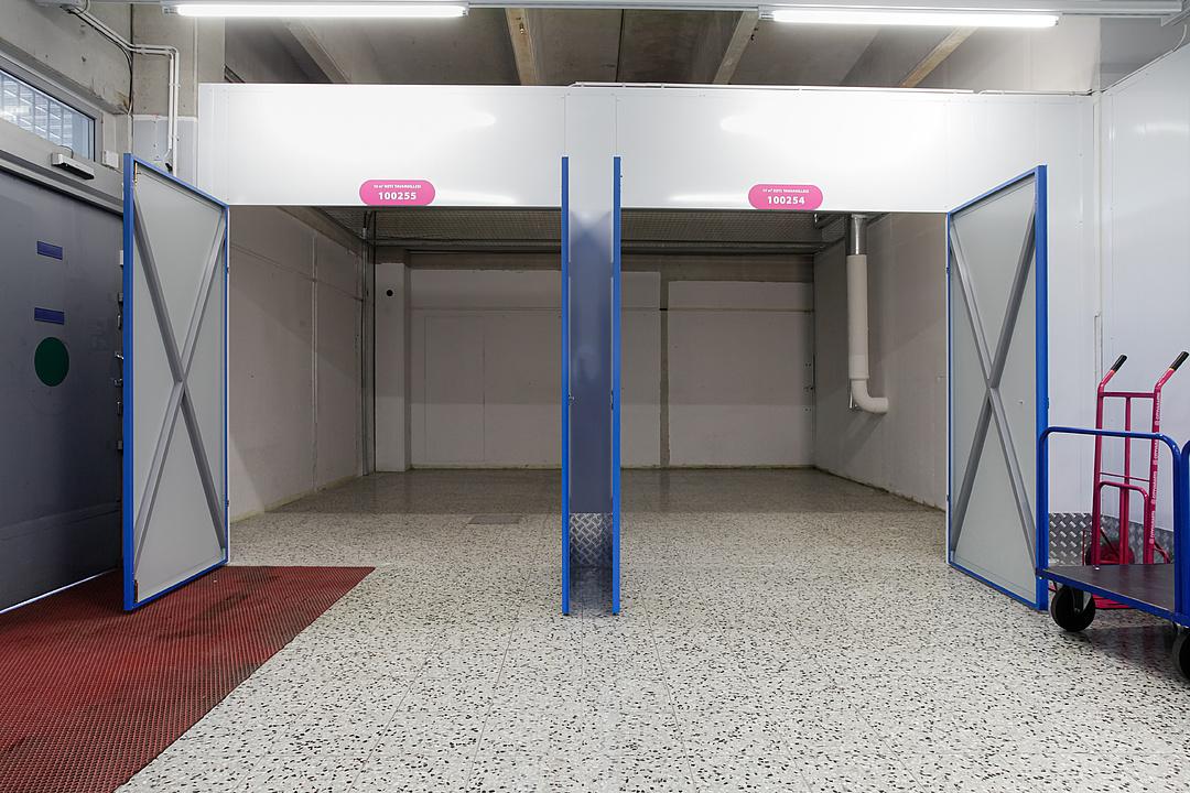 Empty industrial space with two open blue doors, speckled flooring, overhead lights, and a red trolley on the right.