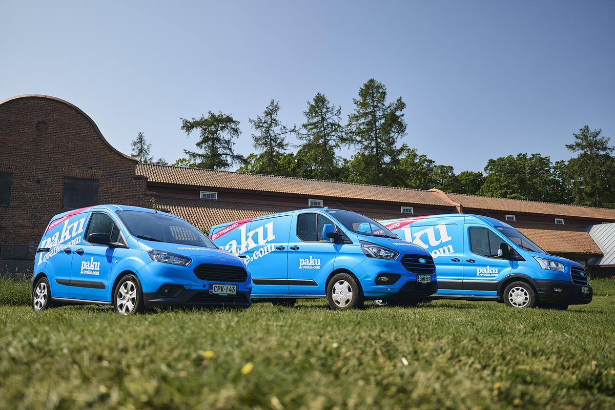 Three blue delivery vans parked on grass in front of a brick building with trees in the background under a clear blue sky.