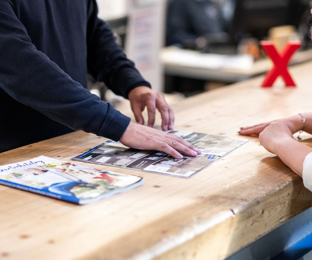 Beratungssituation bei bauXpert: Ein Mitarbeiter zeigt einer Kundin Produktmuster und Kataloge auf einer Holztheke, im Hintergrund steht ein rotes X als Markenzeichen.