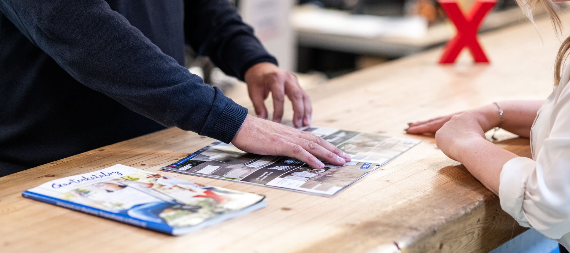 Beratungssituation bei bauXpert: Ein Mitarbeiter zeigt einer Kundin Produktmuster und Kataloge auf einer Holztheke, im Hintergrund steht ein rotes X als Markenzeichen.