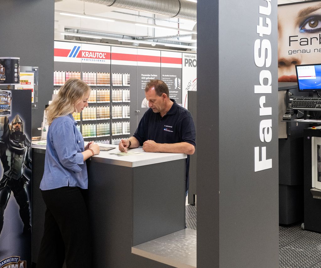 Ein Mitarbeiter zeigt einer Kundin verschiedene Farbmusterkarten zur Abstimmung des gewünschten Farbtons.