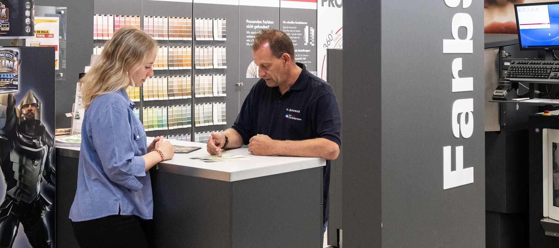 Ein Mitarbeiter zeigt einer Kundin verschiedene Farbmusterkarten zur Abstimmung des gewünschten Farbtons.