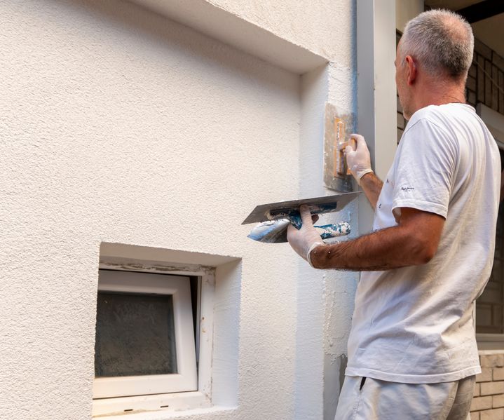 Ein Handwerker verputzt eine weiße Hausfassade mit einer Kelle, neben einem kleinen Kellerfenster.