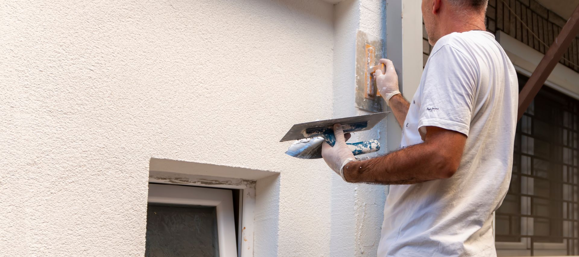 Handwerker verputzt helle Außenfassade neben kleinem Fenster mit Glättkelle – frischer Fassadenputz