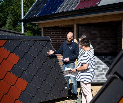 Fachberater erklärt einem Paar verschiedene Dachziegeltypen an einem Musteraufbau im Außenbereich einer Ausstellung für den Gartenbereich..
