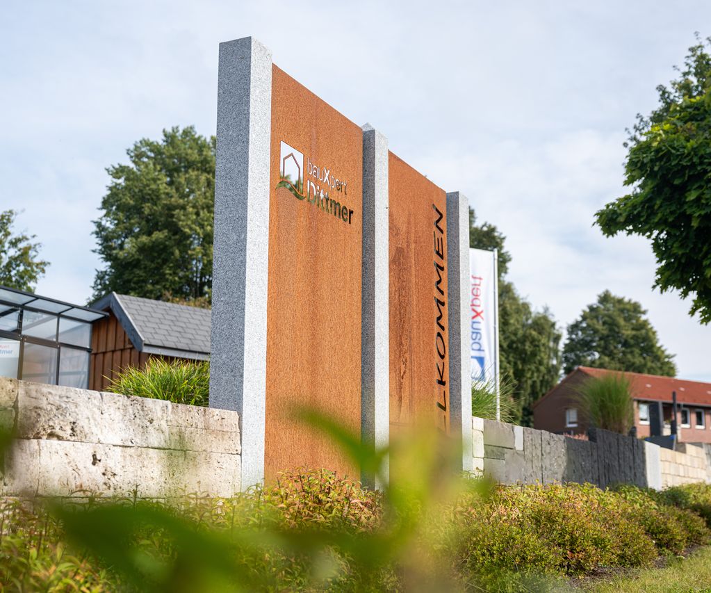 Willkommensschild von bauXpert Dittmer auf dem begrünten Außengelände, mit Flaggen und Glas-Pavillon im Hintergrund