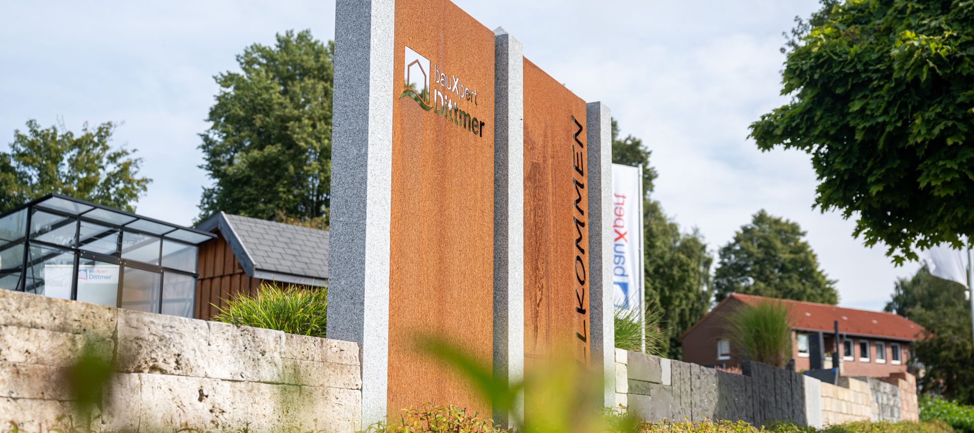 Willkommensschild von bauXpert Dittmer auf dem begrünten Außengelände, mit Flaggen und Glas-Pavillon im Hintergrund