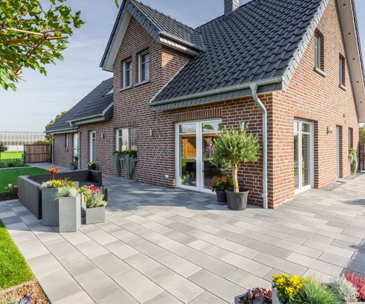 Großformatige helle Pflastersteine auf einer Terrasse vor einem roten Klinkerhaus mit Gartenbereich