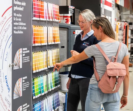 Kundin und bauXpert-Mitarbeiterin wählen gemeinsam eine Farbe an der Farbfächerwand im Fachmarkt aus