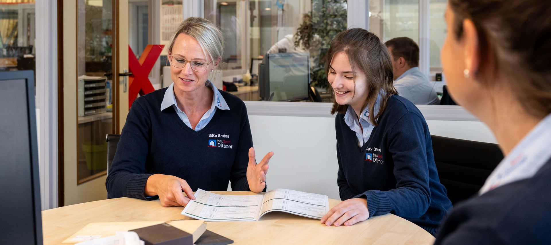 Zwei Mitarbeiterinnen von bauXpert Dittmer im Gespräch mit einer Auszubildenden an einem Besprechungstisch, im Hintergrund ein rotes Deko-X und Bürosituation