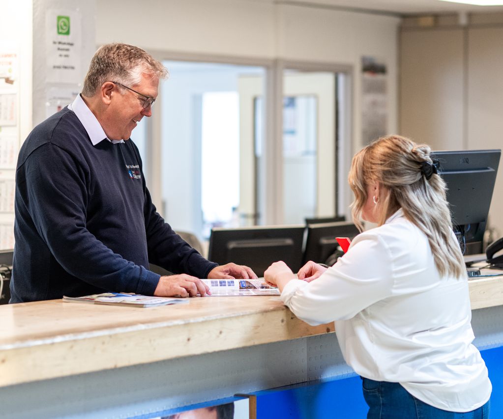 Beratung am Tresen bei bauXpert Dittmer: Ein Mitarbeiter zeigt einer Kundin Angebote im Katalog, im Hintergrund ist das Büro sichtbar