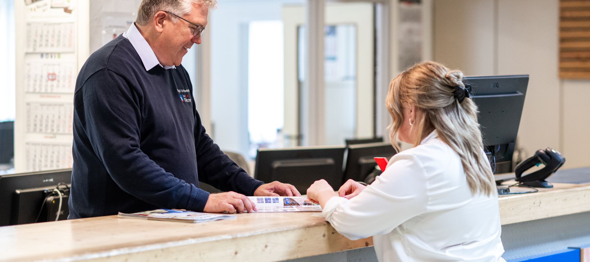 Beratung am Tresen bei bauXpert Dittmer: Ein Mitarbeiter zeigt einer Kundin Angebote im Katalog, im Hintergrund ist das Büro sichtbar
