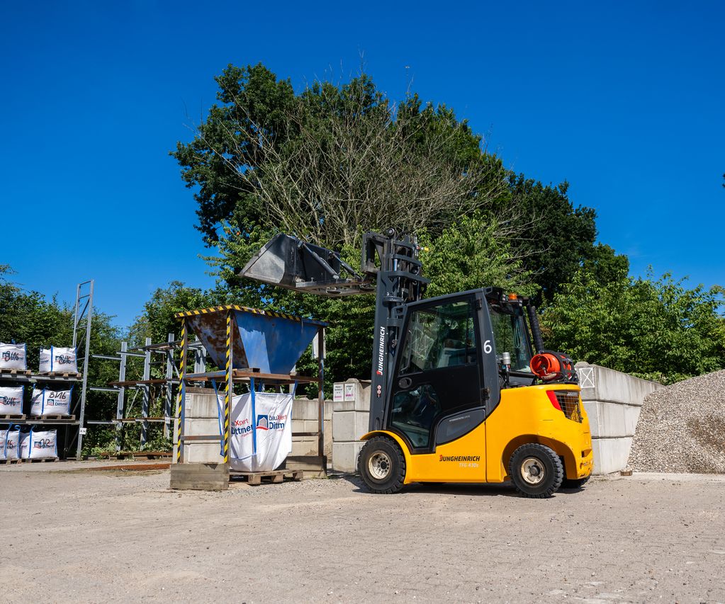 Ein gelber Radlader befüllt einen Metalltrichter, unter dem Big Bags mit dem Logo von bauXpert Dittmer in Lütjenburg stehen. Im Hintergrund sind Kieshaufen und Lagerregale zu sehen.
