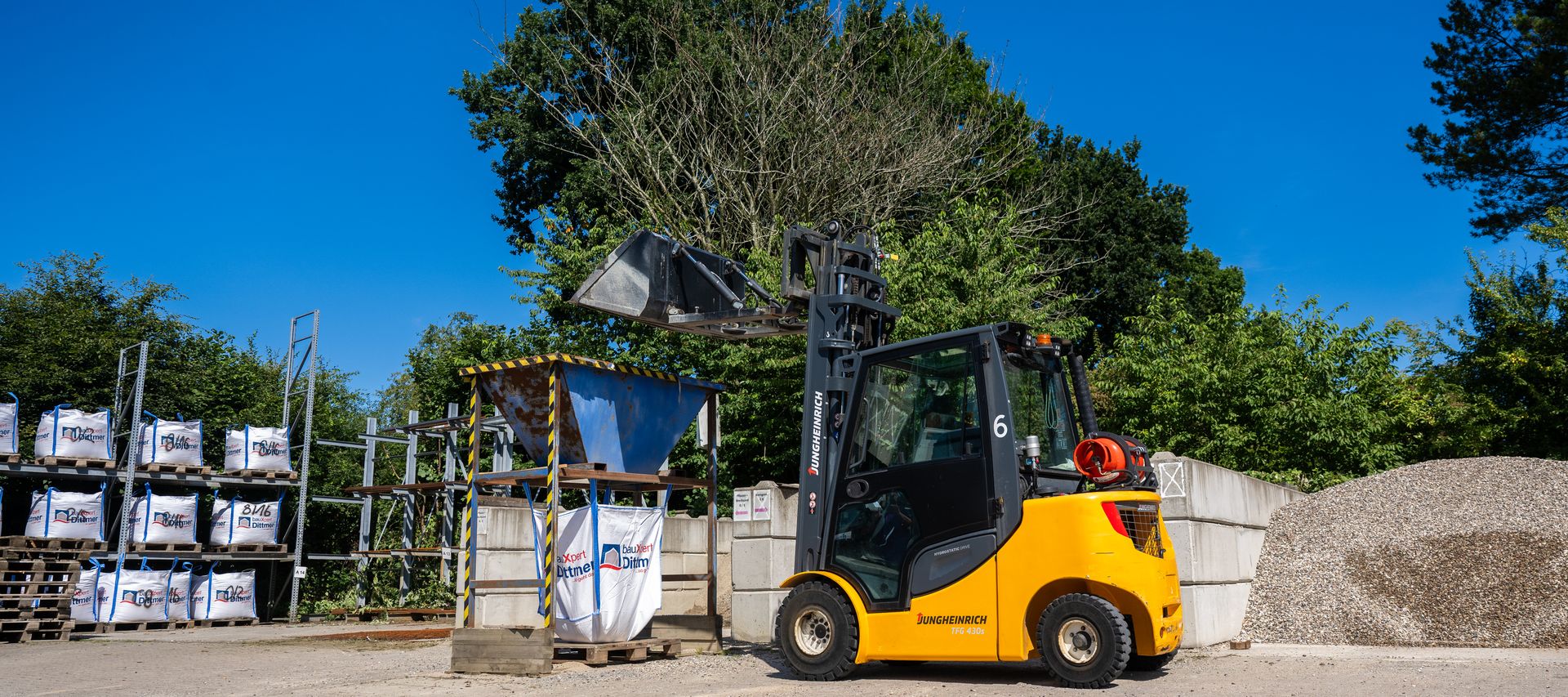 Ein gelber Radlader befüllt einen Metalltrichter, unter dem Big Bags mit dem Logo von bauXpert Dittmer in Lütjenburg stehen. Im Hintergrund sind Kieshaufen und Lagerregale zu sehen.