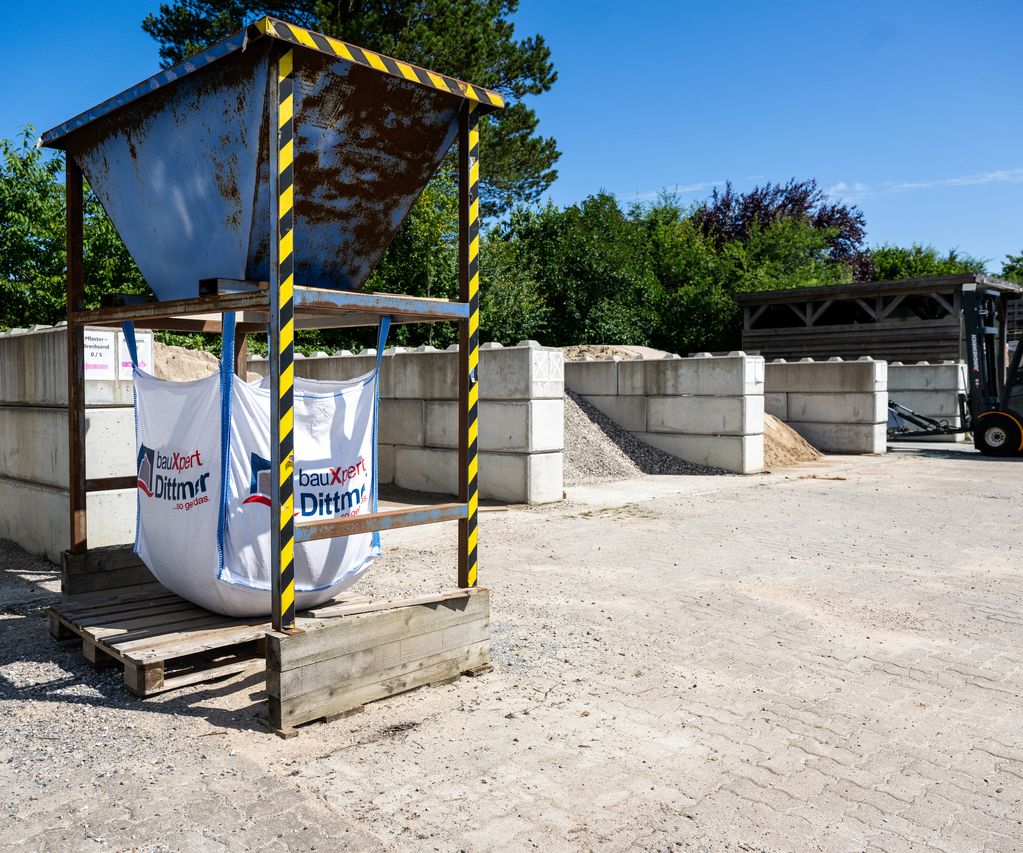 Big-Bag-Station mit zwei befüllten Säcken von bauXpert Dittmer in Lütjenburg, daneben Lagerboxen mit Sand, Kies und einem gelben Radlader im Hintergrund.