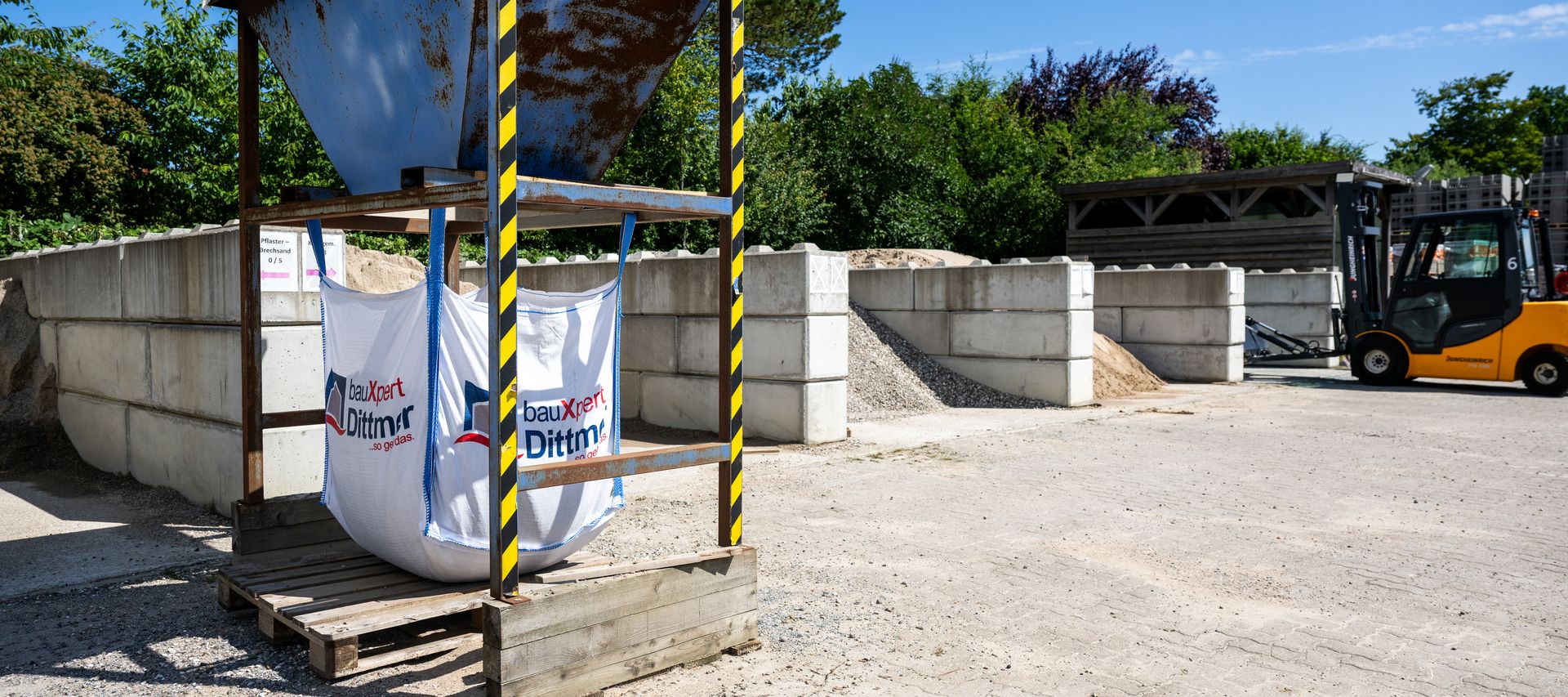 Big-Bag-Station mit zwei befüllten Säcken von bauXpert Dittmer in Lütjenburg, daneben Lagerboxen mit Sand, Kies und einem gelben Radlader im Hintergrund.