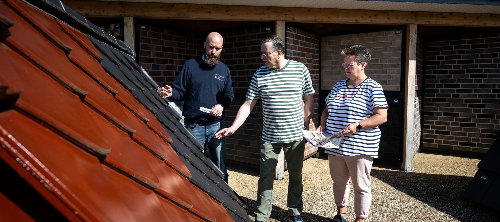 Beratung am Musterdach: Fachberater erklärt rote und anthrazitfarbene Dachziegel – zwei Kund:innen prüfen Farbe und Profil bei bauXpert Dittmer in der GaLa Ausstellung