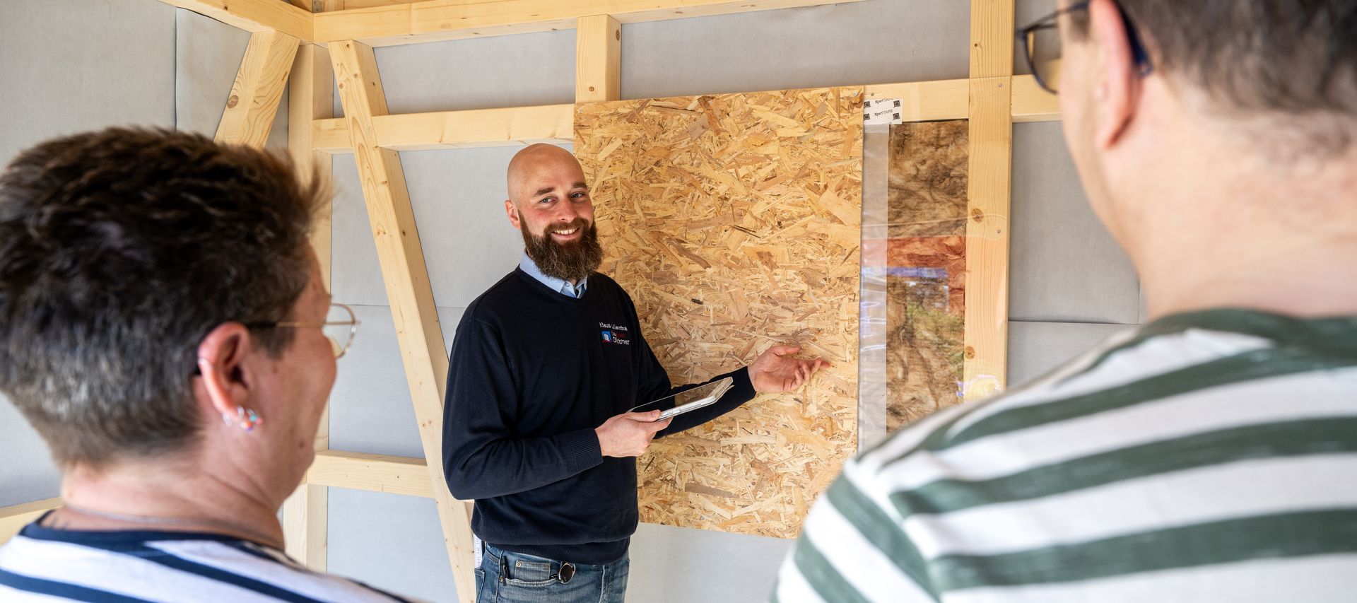 Fachberater erklärt Kunden den Wandaufbau im Holzrahmenbau: OSB-Platte, Dämmung und Dampfbremse am Musterexponat