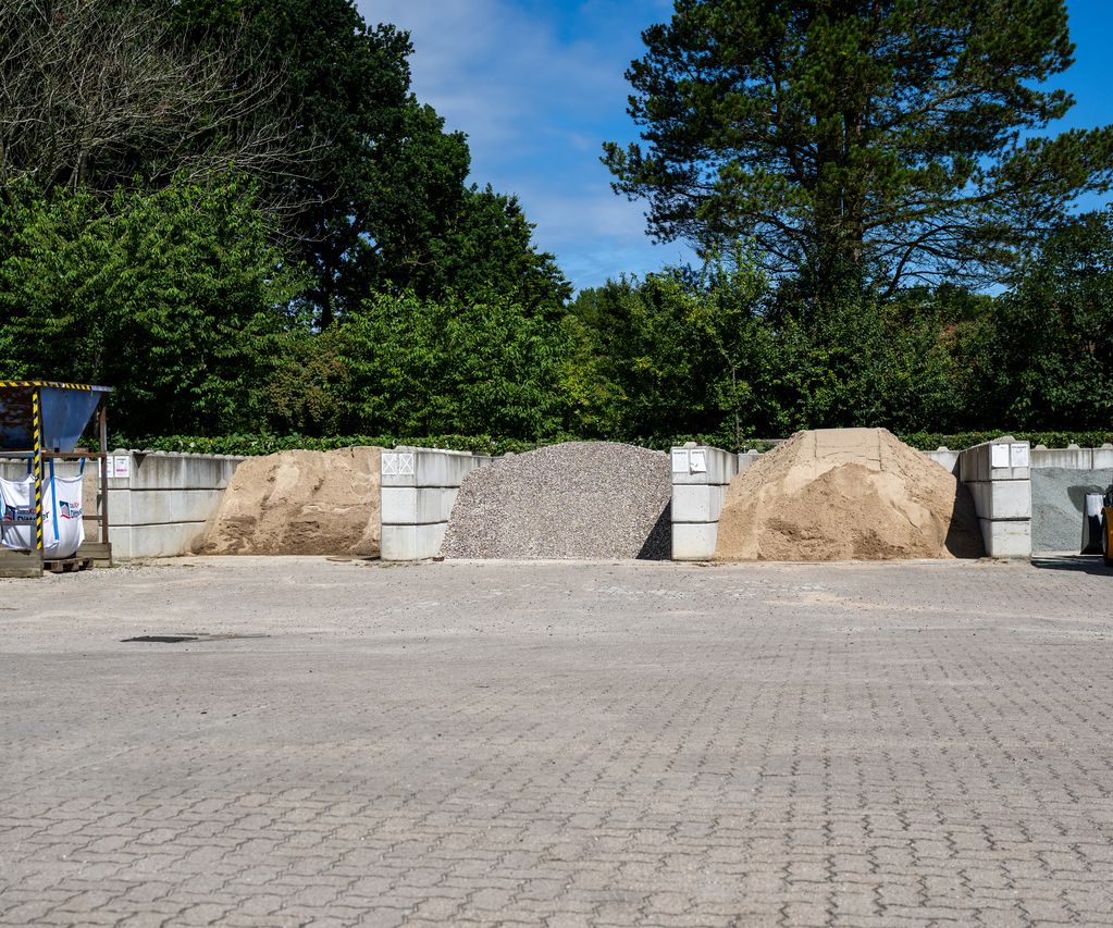 Übersicht des Lagerplatzes bei bauXpert Dittmer in Lütjenburg mit aufgeschüttetem Kies, Sand und Splitt, einem gelben Radlader und einer Big-Bag-Station.