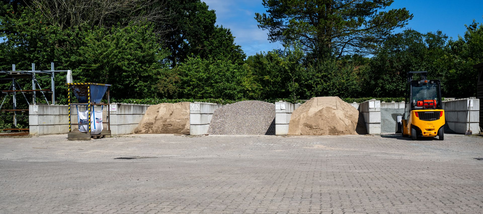 Übersicht des Lagerplatzes bei bauXpert Dittmer in Lütjenburg mit aufgeschüttetem Kies, Sand und Splitt, einem gelben Radlader und einer Big-Bag-Station.