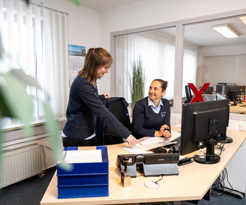 Zwei Mitarbeiterinnen von bauXpert Dittmer in Lütjenburg im Gespräch am Schreibtisch, eine zeigt auf Unterlagen, die andere schaut sie lächelnd an.