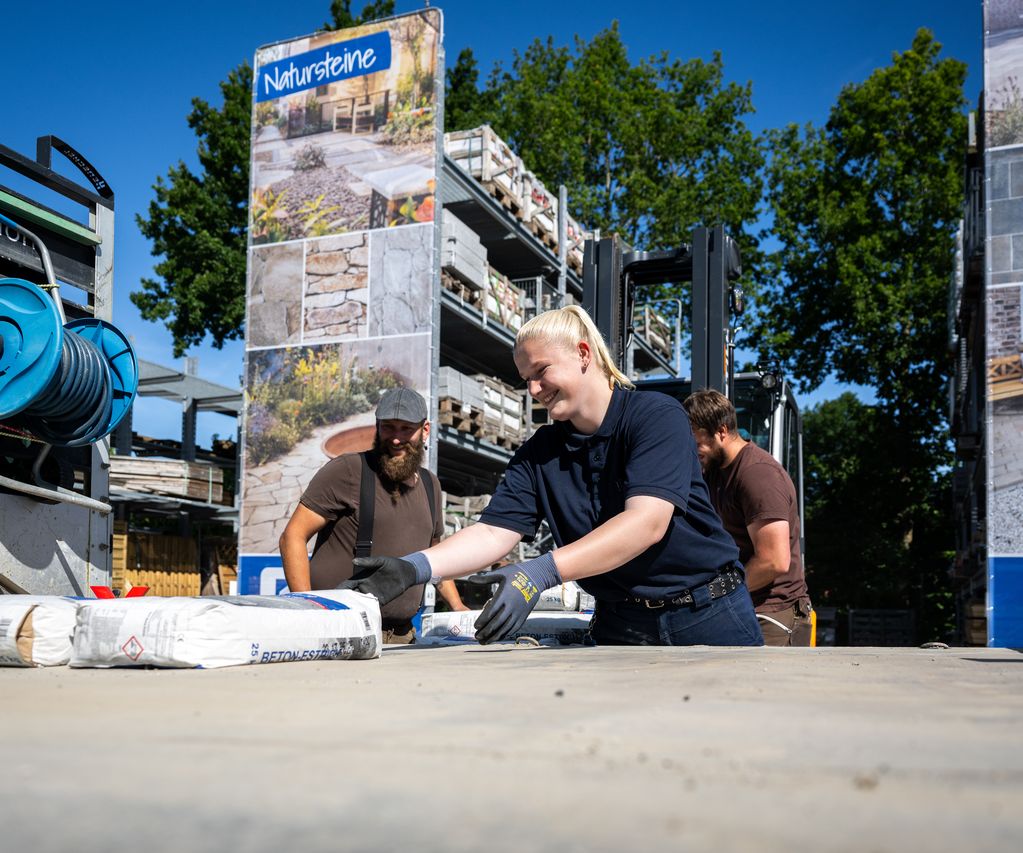 Drei Mitarbeitende von bauXpert Dittmer heben gemeinsam Säcke mit Betonestrich auf einem Lagerplatz, im Hintergrund sind Natursteinpaletten und großformatige Ausstellungstafeln zu sehen.