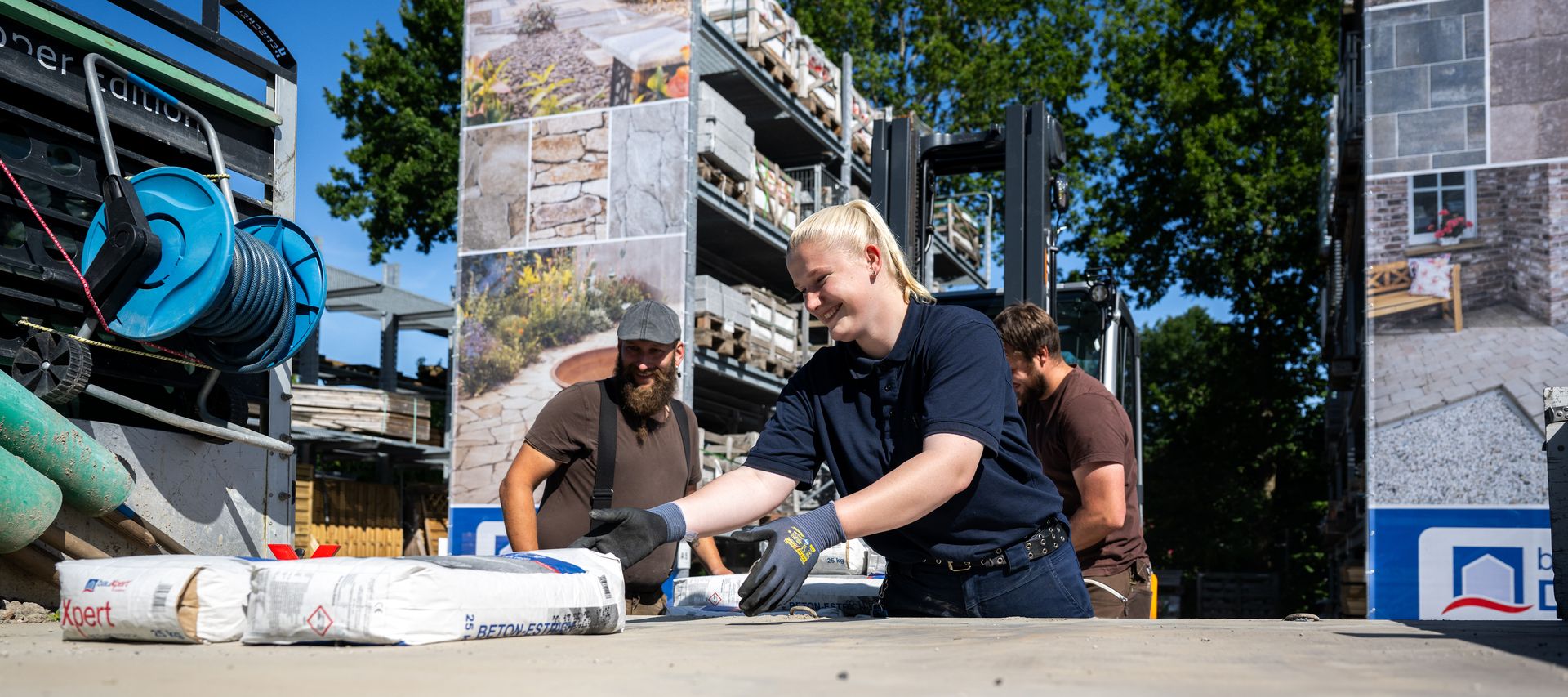 Drei Mitarbeitende von bauXpert Dittmer heben gemeinsam Säcke mit Betonestrich auf einem Lagerplatz, im Hintergrund sind Natursteinpaletten und großformatige Ausstellungstafeln zu sehen.