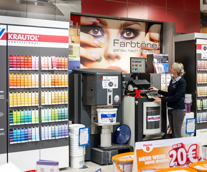 Mitarbeiterin von bauXpert Dittmer bedient die Farbmischanlage im Fachmarkt vor einer großen Wand mit Farbkarten