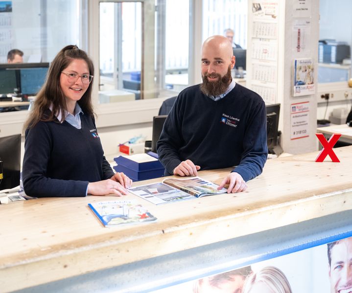 Zwei Mitarbeitende von bauXpert Dittmer stehen am Tresen und lächeln in die Kamera, aufgeschlagene Kataloge liegen vor ihnen