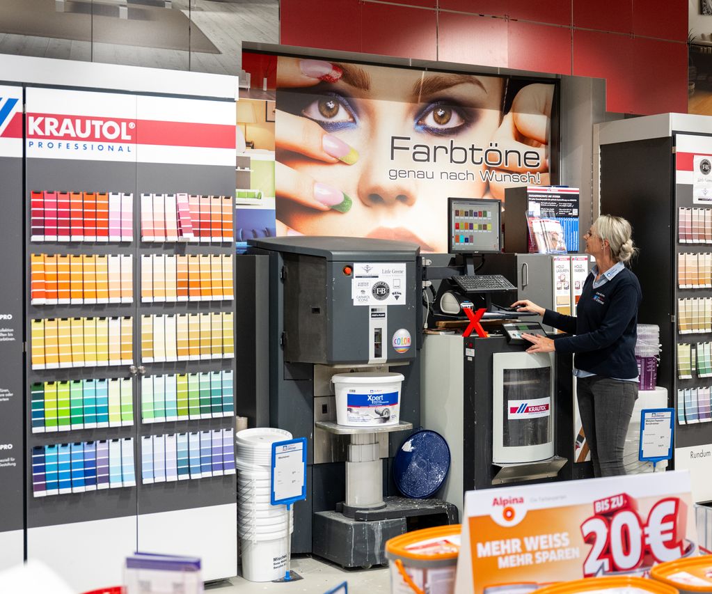 Eine Mitarbeiterin von bauXpert Dittmer steht an der Farbmischanlage im Fachmarkt und bedient den Computer. Um sie herum befinden sich Farbtonkarten und Eimer mit weißer Farbe. Im Hintergrund ist ein großes Bild mit dem Schriftzug „Farbtöne genau nach Wunsch!“ zu sehen.