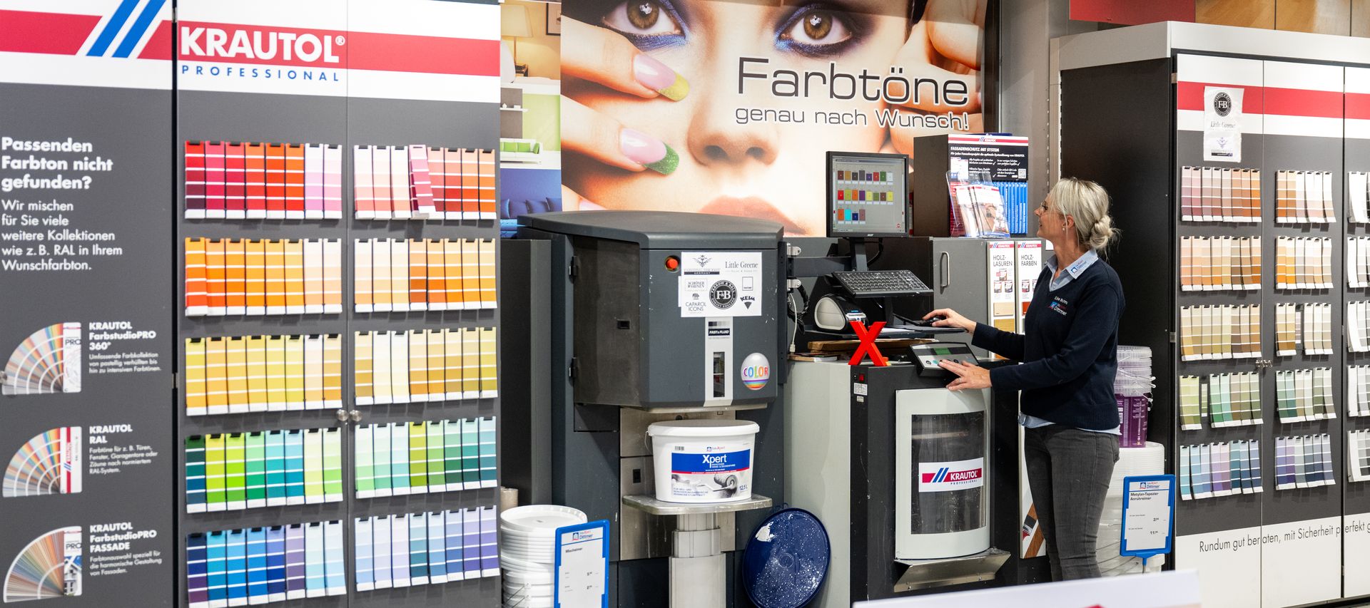 Eine Mitarbeiterin von bauXpert Dittmer steht an der Farbmischanlage im Fachmarkt und bedient den Computer. Um sie herum befinden sich Farbtonkarten und Eimer mit weißer Farbe. Im Hintergrund ist ein großes Bild mit dem Schriftzug „Farbtöne genau nach Wunsch!“ zu sehen.
