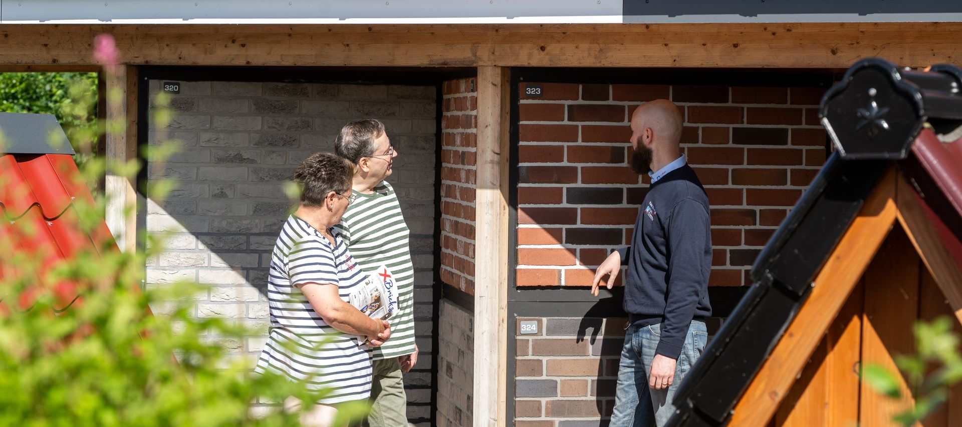 Fachberater erklärt einem Paar unterschiedliche Klinkerverblender an der Musterwand im Außenbereich von bauXpert Dittmer