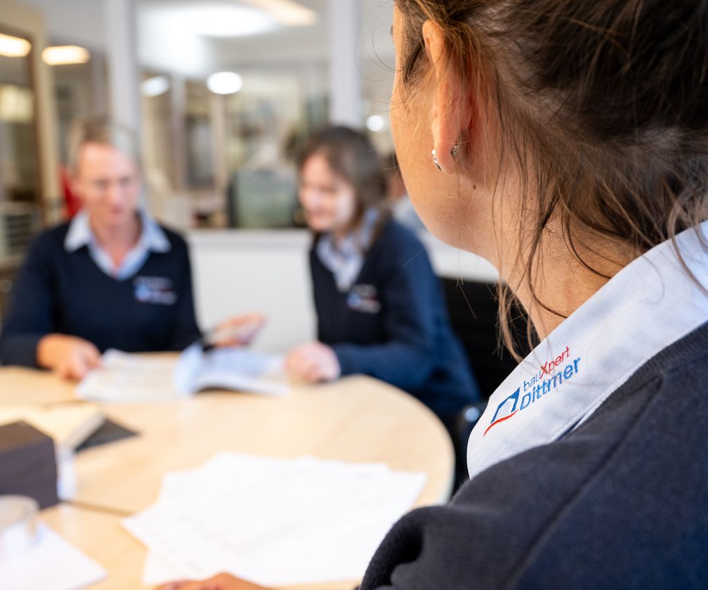 Nahaufnahme einer Mitarbeiterin von bauXpert Dittmer in Lütjenburg mit sichtbarem Logo auf dem Hemdkragen, im Hintergrund Kolleginnen im Gespräch bei einem Teammeeting.