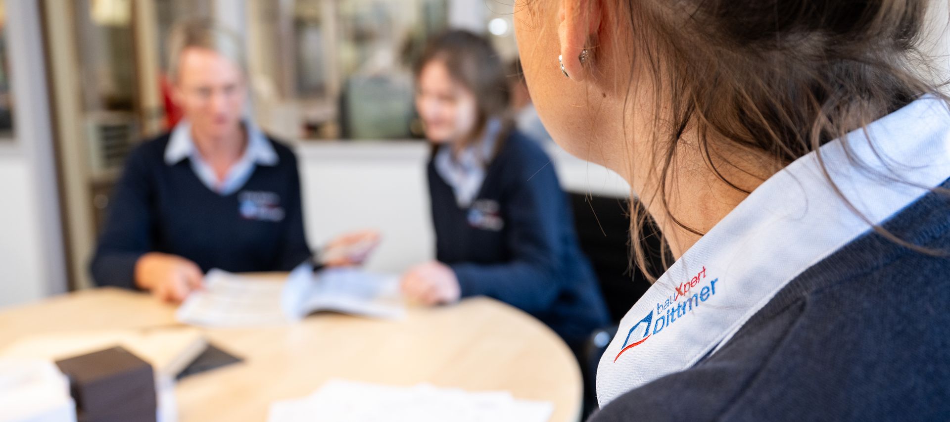 Nahaufnahme einer Mitarbeiterin von bauXpert Dittmer in Lütjenburg mit sichtbarem Logo auf dem Hemdkragen, im Hintergrund Kolleginnen im Gespräch bei einem Teammeeting.