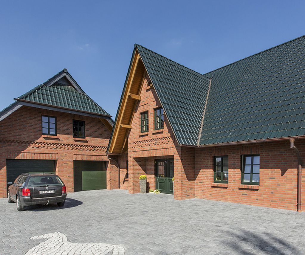 A large brick house with green roofs, adjacent garage, and a parked car on a paved driveway under a clear blue sky.