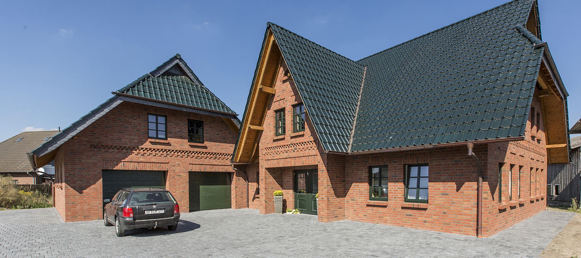 A large brick house with green roofs, adjacent garage, and a parked car on a paved driveway under a clear blue sky.