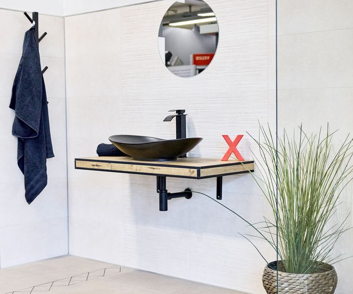 Fliesenausstellung: Modernes Badezimmer mit schwarzem Aufsatzwaschbecken, runden Spiegel, Topfpflanze und Handtuch an der Wand, rotes ‚X‘ markiert den Waschbereich