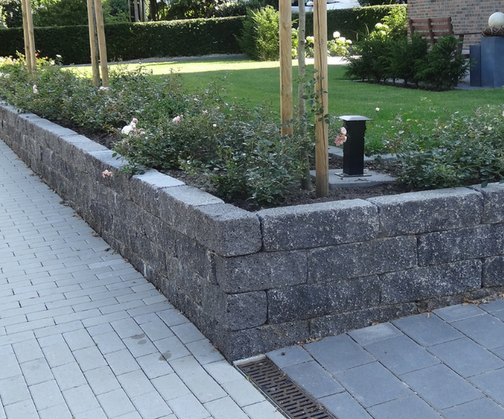 Trockenmauer mit kleinen Sträuchern und jungen Bäumen entlang eines gepflasterten Weges, Hofeinfahrt eines modernen Backsteingebäudes.