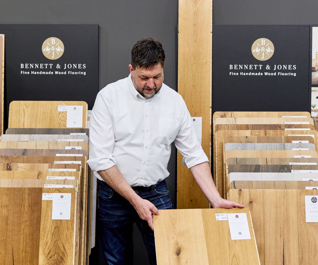 Ein Verkäufer begutachtet eine Parkett Holzfußbodenprobe in einem Ausstellungsraum, in dem um ihn herum verschiedene Parkettböden zum Verkauf ausgestellt sind.