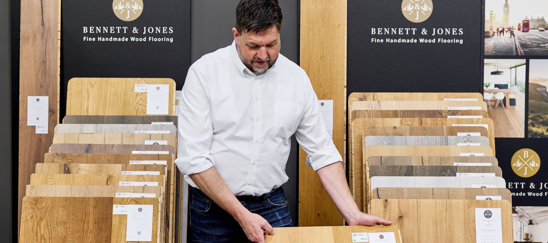 Ein Verkäufer begutachtet eine Parkett Holzfußbodenprobe in einem Ausstellungsraum, in dem um ihn herum verschiedene Parkettböden zum Verkauf ausgestellt sind.