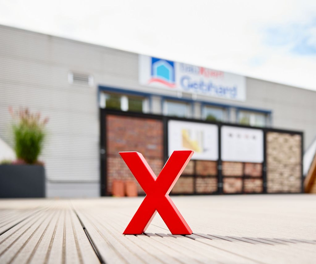 Rotes X-Logo auf einer Terrasse vor unscharfer Außenausstellung mit Verblendsteinen und Dachmustern