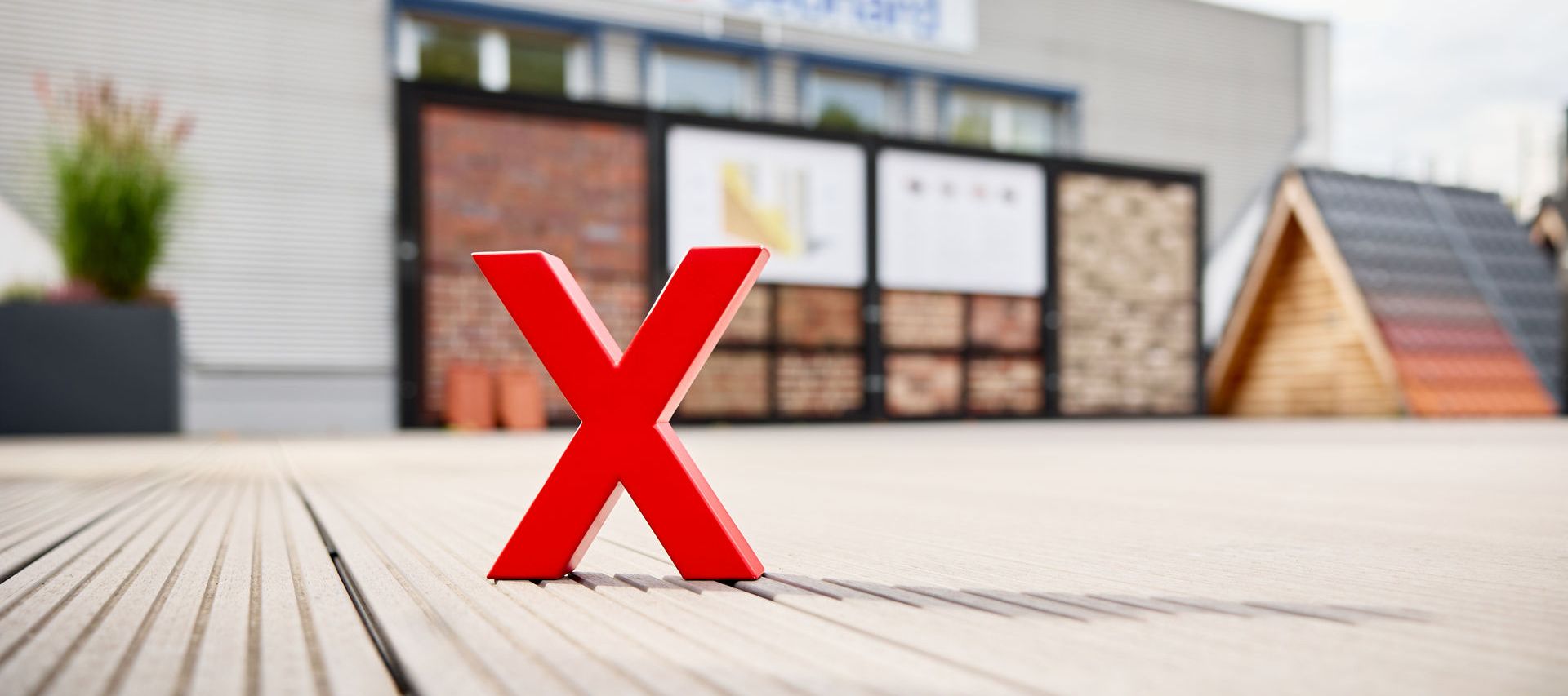Rotes X-Logo auf einer Terrasse vor unscharfer Außenausstellung mit Verblendsteinen und Dachmustern