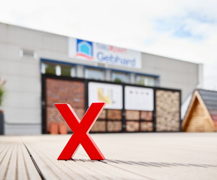Rotes X-Logo auf einer Terrasse vor unscharfer Außenausstellung mit Verblendsteinen und Dachmustern