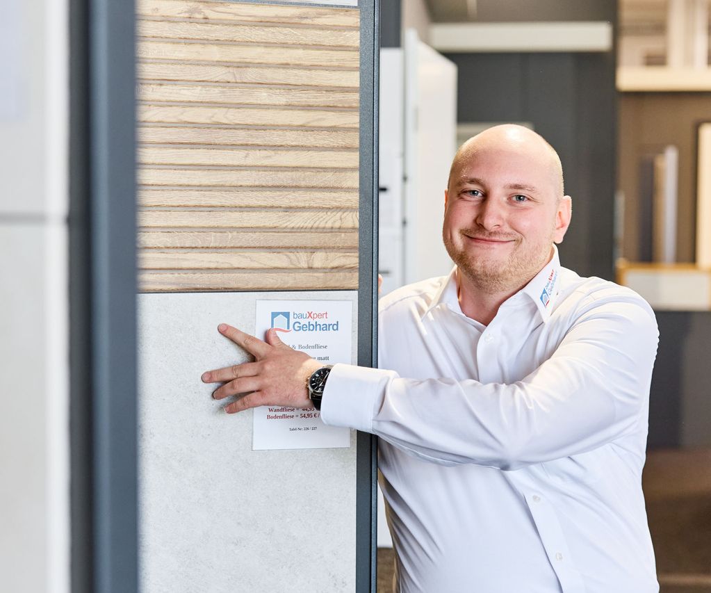 Ein Fliesen Fachberater in weißem Hemd lächelt, während er sich an einem Fliesendisplay in einer Fliesenausstellung anlehnt.