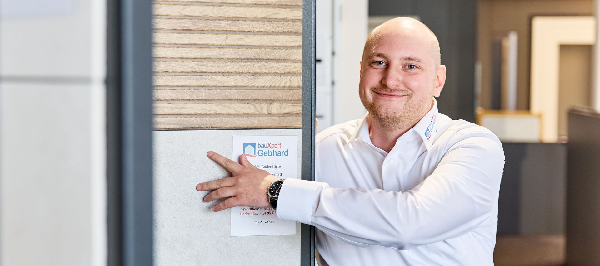 Ein Fliesen Fachberater in weißem Hemd lächelt, während er sich an einem Fliesendisplay in einer Fliesenausstellung anlehnt.