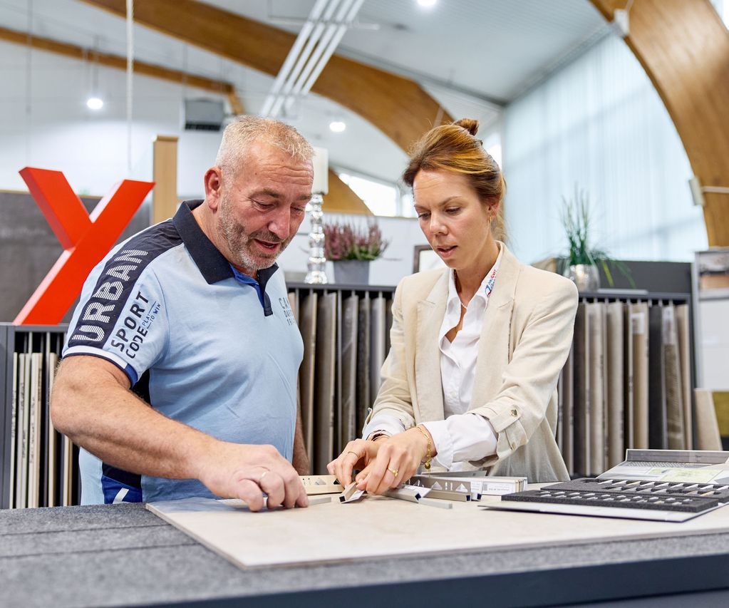 A man and a woman discuss tile samples in a showroom, surrounded by various flooring options and a large red "X" in the background.