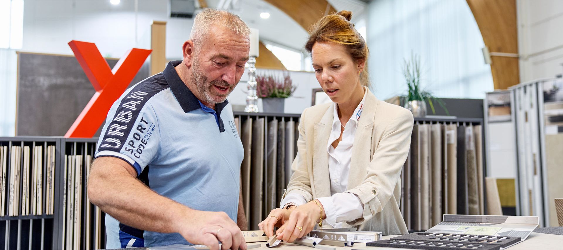 A man and a woman discuss tile samples in a showroom, surrounded by various flooring options and a large red "X" in the background.
