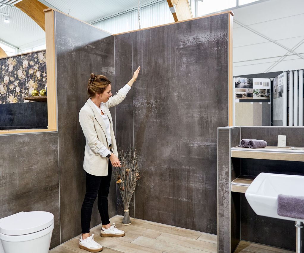 Junge Frau zeigt in eine Fliesenausstellung auf ein modernes Badezimmer-Setup mit schwarzen Fliesen, Toilette und Waschbecken.
