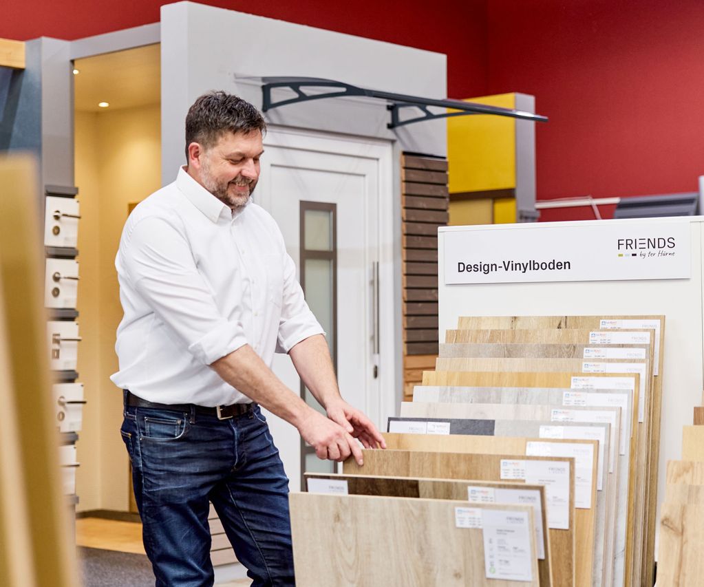 Ein Verkäufer in einem weißen Hemd begutachtet Vinylbodenbelagsmuster bei bauXpert in Zeven, umgeben von verschiedenen Vinylböden in Holtönen..