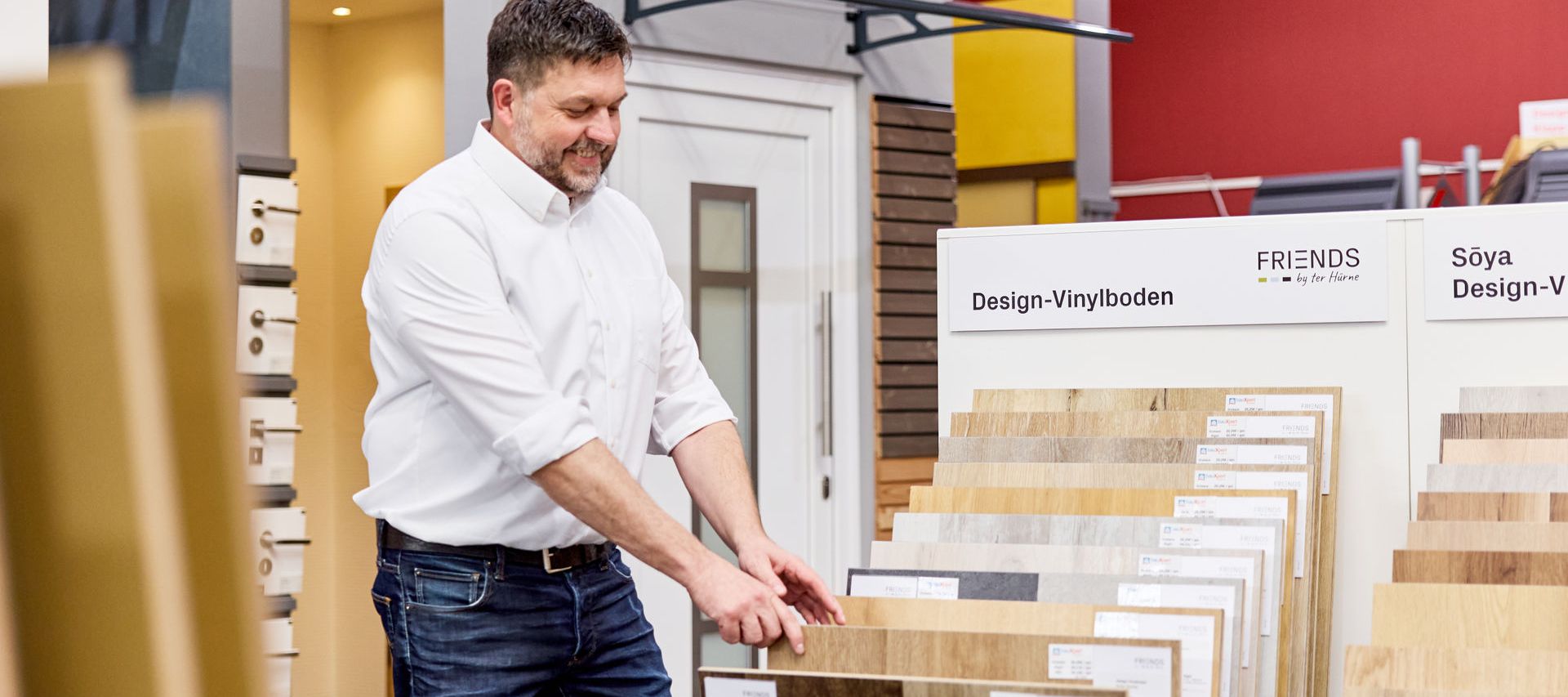 Ein Verkäufer in einem weißen Hemd begutachtet Vinylbodenbelagsmuster bei bauXpert in Zeven, umgeben von verschiedenen Vinylböden in Holtönen..